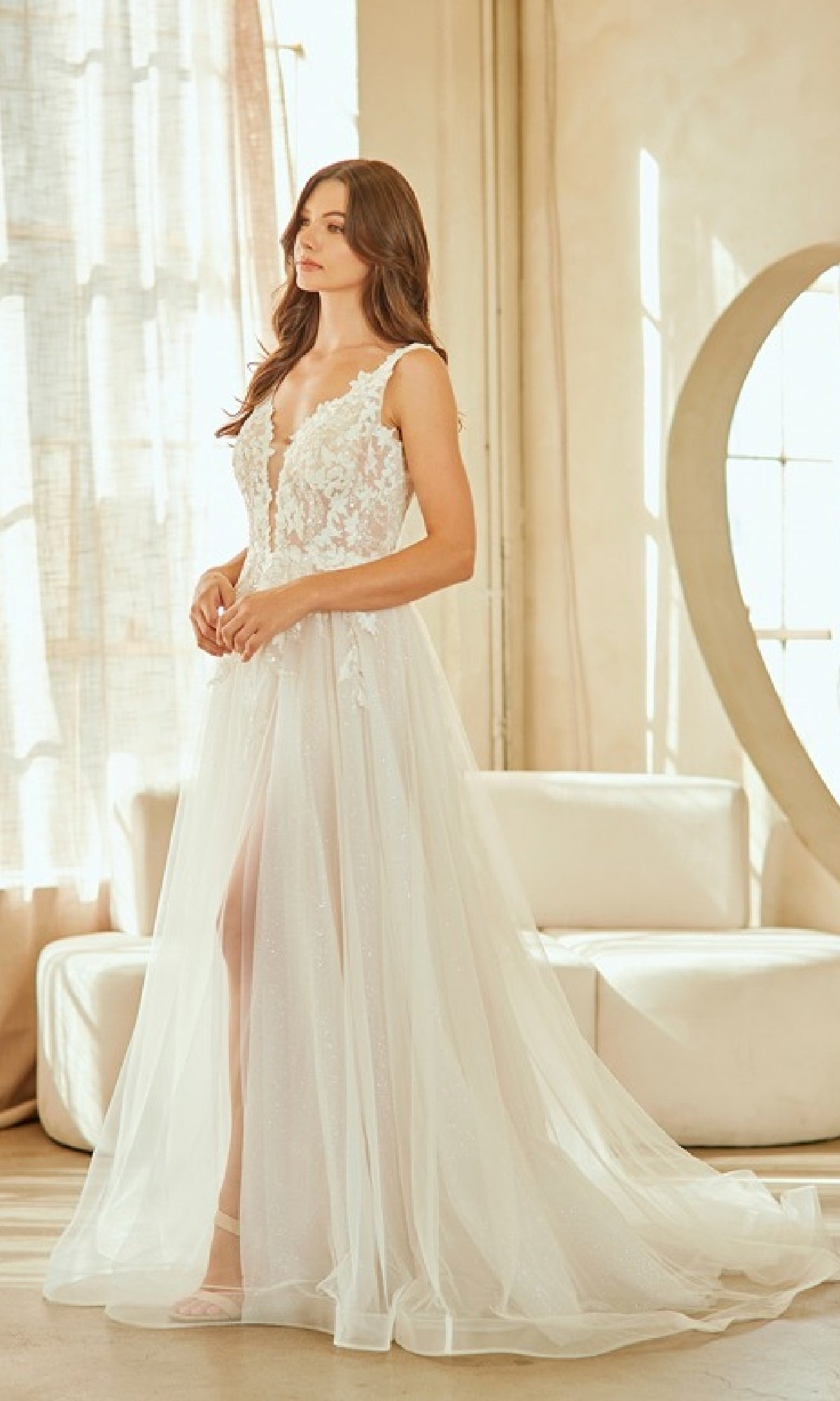 A woman stands indoors wearing the Amelia Couture 2140 long wedding dress, a sleeveless gown with lace embroidery, a deep V neckline, and a sheer tulle overlay.